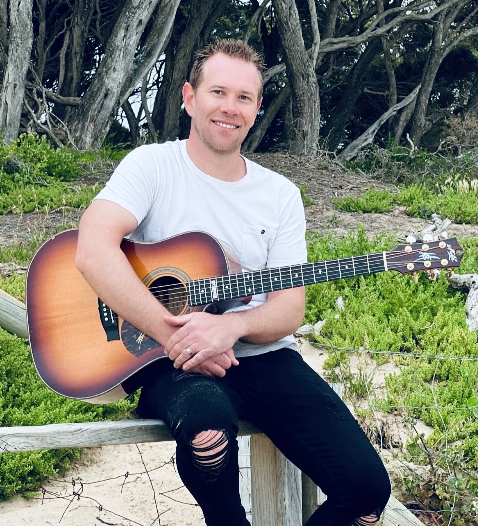 Man in white tshirt sits on a low wall with a guitar