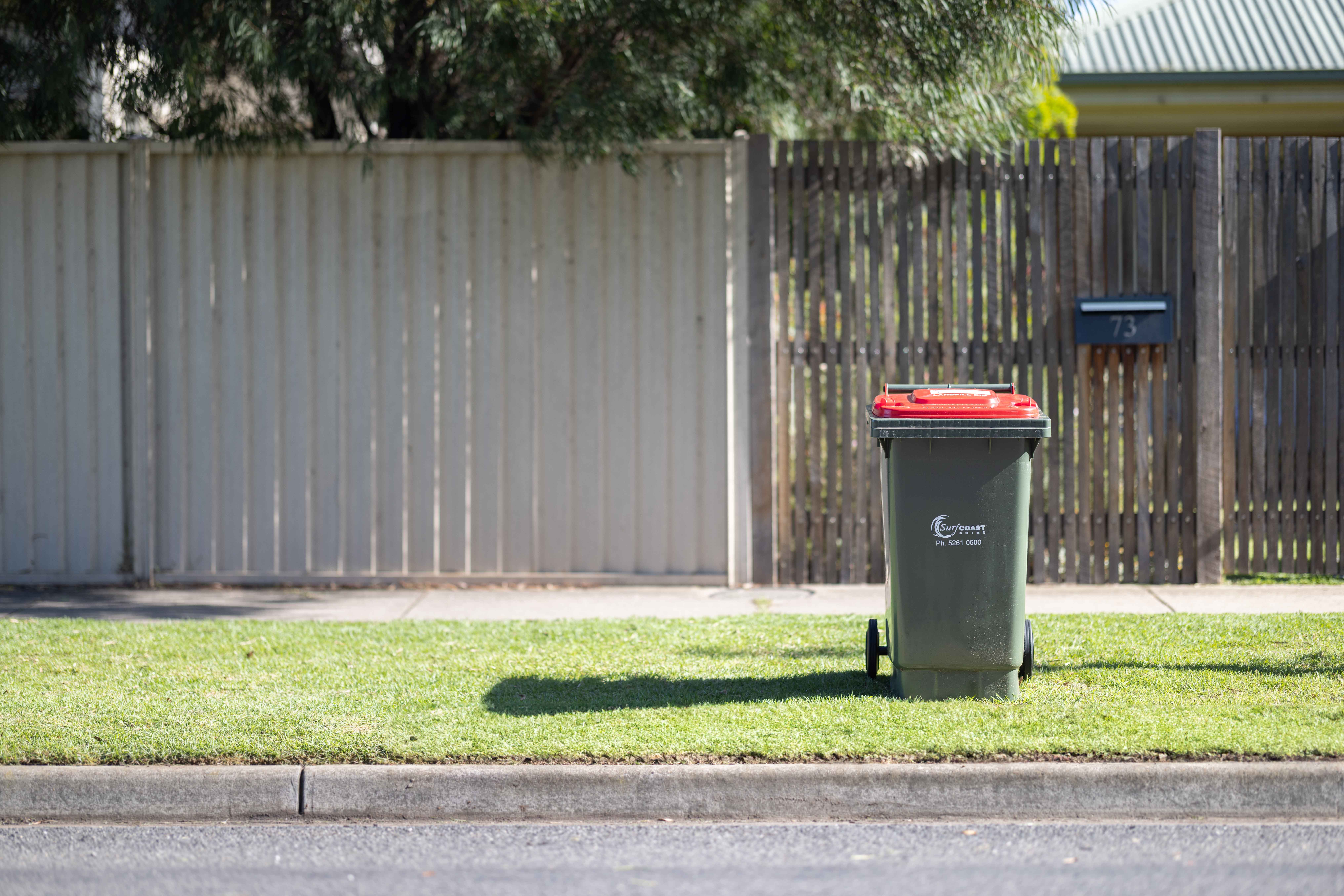 Red lidded landfill bin sitting on a kerb in front of a fence