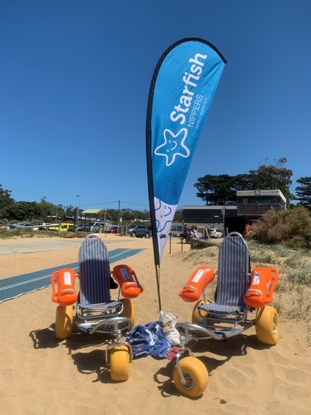 Two beach wheelchairs parked in front of a Starfish Nippers sign