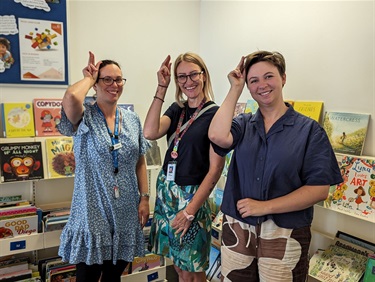 Librarian Frances with Coordinator of Child and Family Health Amy Clemens, and Auslan interpreter, standing in front of bookshelves, signing 'Library' in Auslan, at the Torquay Library.